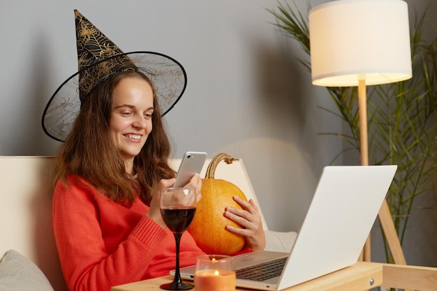 Image of satisfied delighted woman wearing jumper and witch hat sitting on sofa in front of laptop and using mobile phone for online communication with friends during Halloween holiday