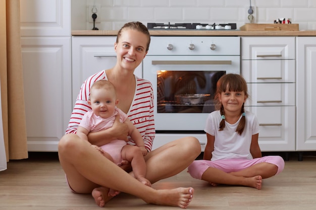 Изображение довольной счастливой улыбающейся женщины в полосатой рубашке, сидящей на полу на кухне со своими маленькими дочерьми, мать которых держит младенца, люди смотрят в камеру с оптимизмом