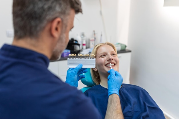 Image of satisfied young woman sitting in dental chair at medical center while professional doctor fixing her teeth Female dentist choosing filling shade for smiling woman using tooth scale sample