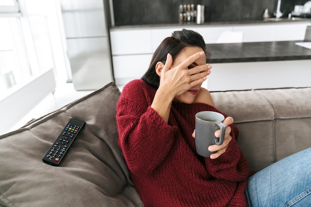 目を覆っているコーヒーを飲んでいるソファ時計テレビの家の屋内で怖がっている感情的な女性の画像。