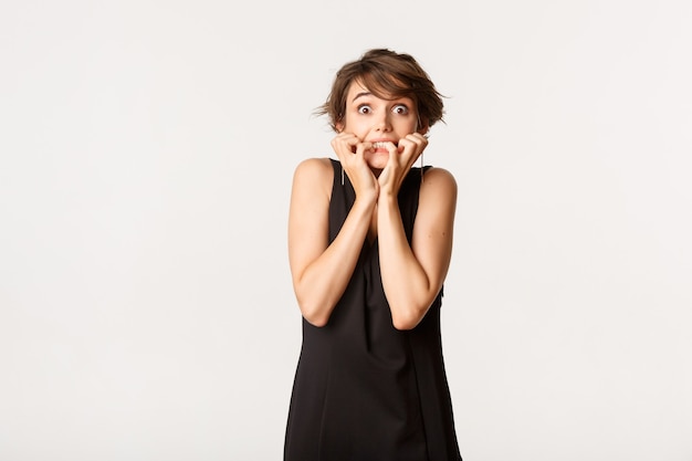 Image of scared and insecure brunette woman in black dress, trembling from fear and biting fingernails, standing over white.