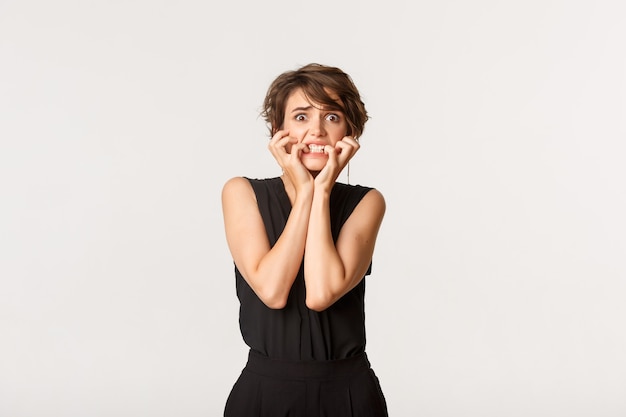 Image of scared woman trembling from fear, looking frightened and biting fingernails, standing white.