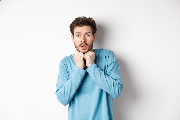 Image of scared young man trembling from fear, holding hands near face and staring at camera startled, standing over white background.