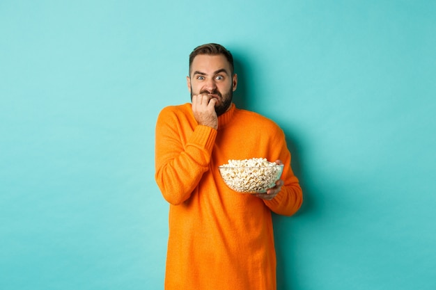 Image of scared young man watching horror movie, biting fist and looking terrified, holding bowl of