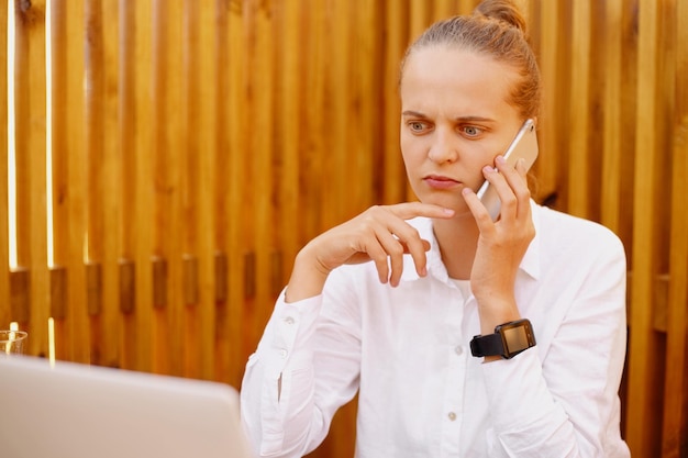 Image of serious angry young adult woman with wearing white shirt talking on smart phone against wooden wall looking away and holding chin having strict bossy expression