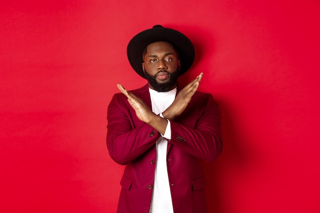 Image of serious Black man strongly disagree, showing cross sign to stop, prohibit something, standing over red background