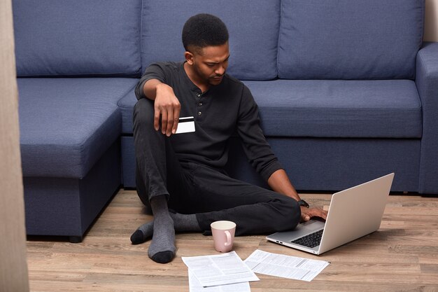 Image of serious pensive Afroamerican man staying at home, being confused with online services, holding credit card