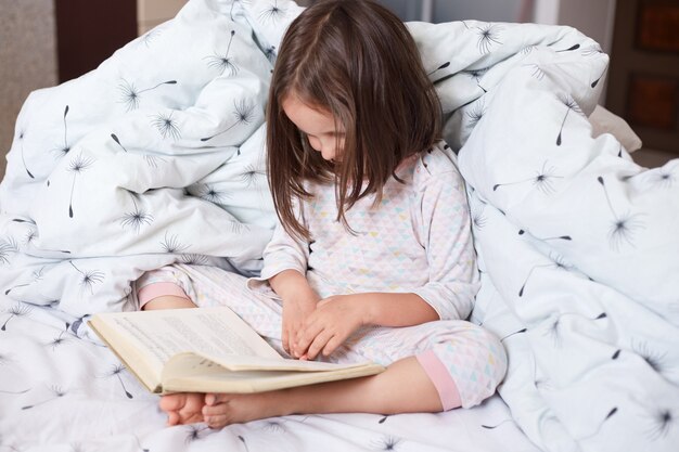 Image of serious preschooler girl reading book in bed