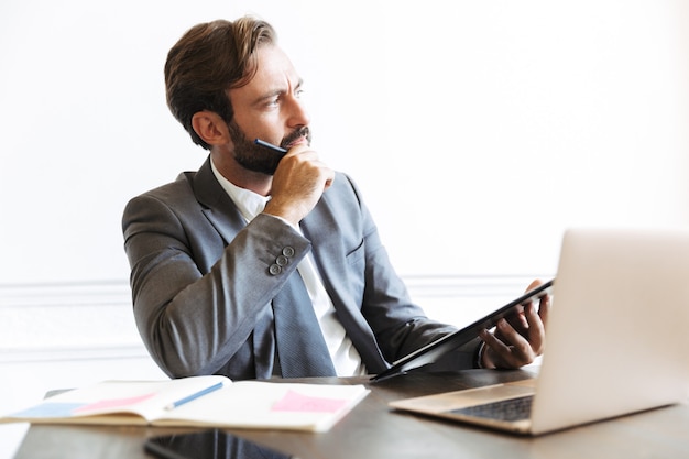 Image of serious puzzled businessman wearing formal suit reading documents while working at laptop in office