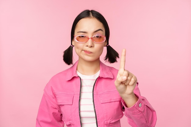 Image of serious stylish asian girl in sunglasses showing stop prohibit gesture taboo sign saying no standing over pink background