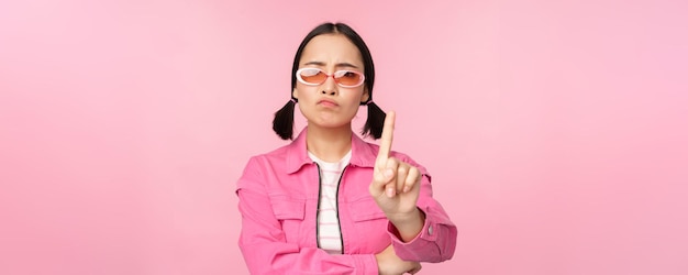 Image of serious stylish asian girl in sunglasses showing stop prohibit gesture taboo sign saying no standing over pink background