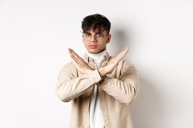 Image of serious young guy in glasses say no, showing cross gesture, make stop sign to disagree or prohibit something, standing on white wall.