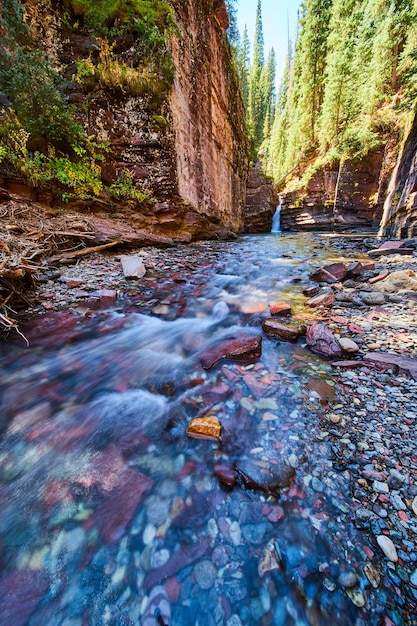 Image of Shallow river with colorful rocks at bottom of canyon and waterfall in distance