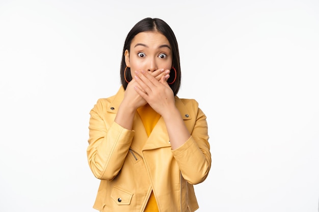 Image of shocked asian girl shuts mouth close lips and looking speechless startled face expression standing over white background