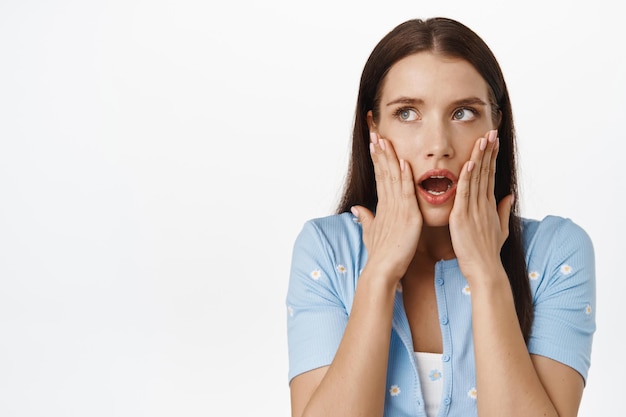 Image of shocked brunette woman gasp, open mouth startled, holding hands on face, staring at upper left corner sale or banner, standing speechless in stupor against white background