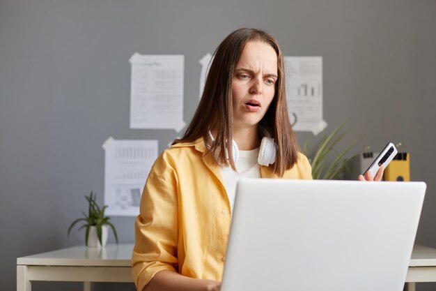 Image of shocked confused Caucasian woman with brown hair working on laptop posing in office holding cell phone in hands and looking at notebook display with big eyes