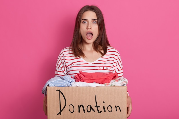 Image of shocked emotional lady with widely opened mouth and eyes, being surprised, having carton box with donated clothes in hands, looking directly at camera. People and volunteering concept.