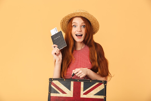 Image of a shocked excited cute young pretty redhead woman posing isolated over yellow wall holding suitcase and passport with tickets