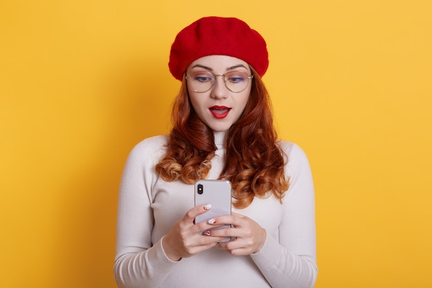 Image of shocked student girl in red beret posing with cellphone in hands on yellow