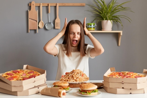 Image of shocked young adult Caucasian woman wearing white casual Tshirt sitting at table in kitchen screaming with astonishment keeps mouth open looking at junk food