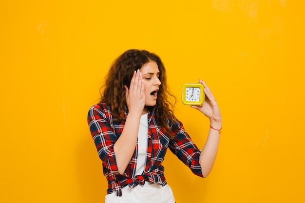 Image shocked young woman  isolated over yellow wall, showing alarm clock