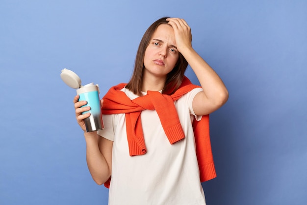 Image of sick tired Caucasian woman wearing white Tshirt and orange sweater tied over shoulders holding thermos with coffee suffering headache standing isolated on blue background