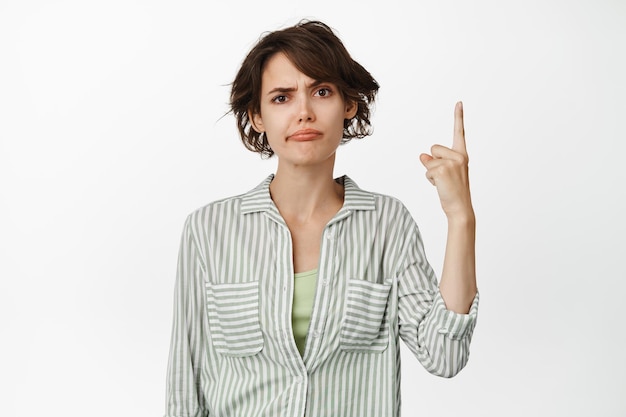 Image of skeptical young woman smirking, frowning upset, pointing finger up with doubtful, unsure face expression, standing over white background