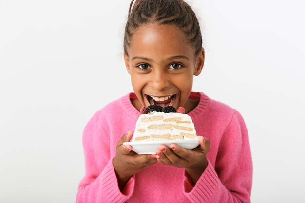 Image of smiling african american girl eating piece of torte while looking at camera isolated over white wall