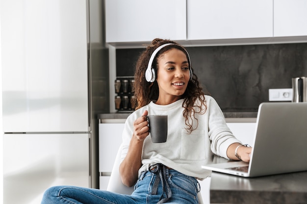 Image of smiling african american girl wearing headphones using laptop, while sitting in bright kitchen