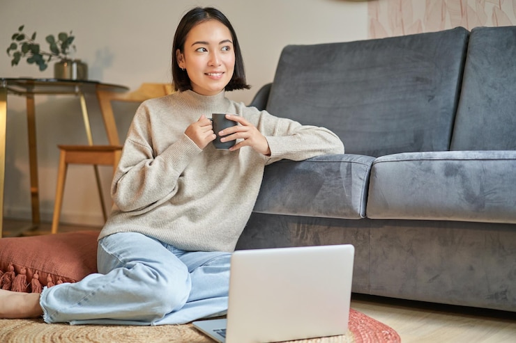 Photo image of smiling asian woman drinking hot tea holding cup and sitting near laptop on floor resting a