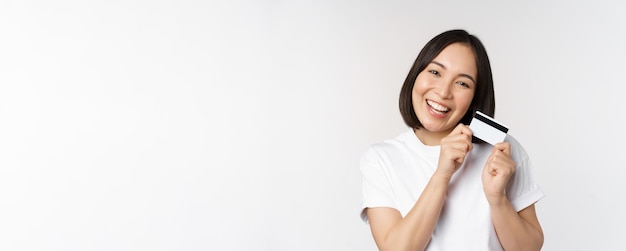 Image of smiling asian woman hugging credit card buying contactless standing in white tshirt over wh