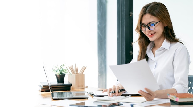 Immagine di una bella donna sorridente che lavora su un documento e un computer portatile mentre è seduta al tavolo in ufficio