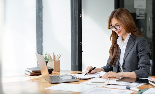 Immagine di una bella donna sorridente che lavora su un documento e un computer portatile mentre è seduta al tavolo in ufficio