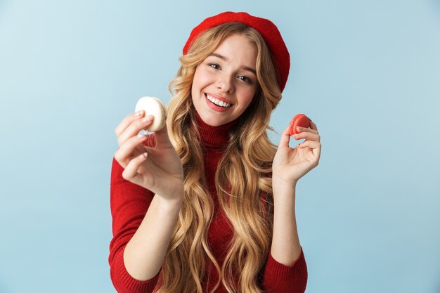 Image of smiling blond woman 20s wearing red beret holding macaron cookies isolated 