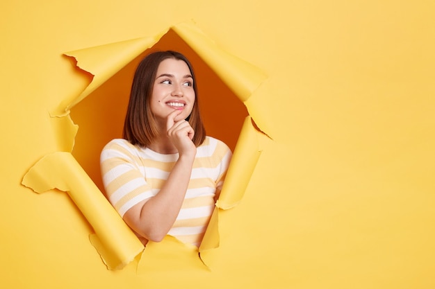 Image of smiling Caucasian woman stands in torn paper hole looking away with dreamy expression looks happy looking through breakthrough of yellow background