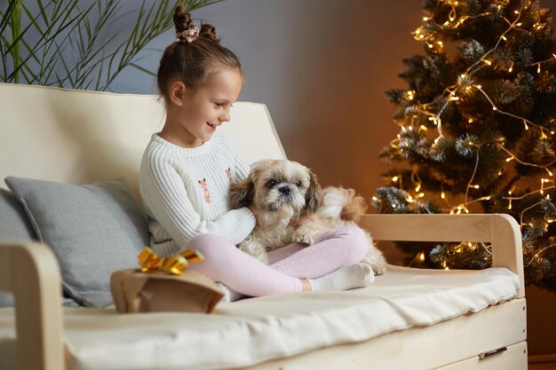 Image of smiling charming little girl with two hair buns wearing casual clothing sitting on sofa with dog expressing positive emotions Christmas celebrations