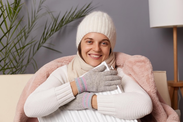 Image of smiling delighted woman in coat and hat sit in cold\
living room with radiator looking at camera embracing heater being\
in good mood despite the lack of heat in her apartment
