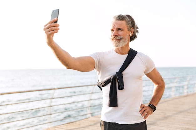 Image of smiling elderly man in sportswear taking selfie photo on cellphone at promenade in morning