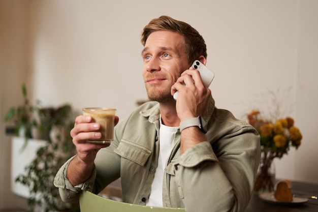 Image of smiling goodlooking young blond man with phone drinking coffee in cafe talking to someone