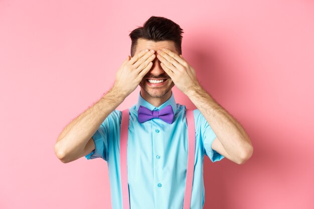 Photo image of smiling handsome guy waiting for surprise with closed eyes, standing in fancy outfit on pink background.