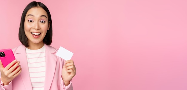 Image of smiling happy asian businesswoman showing credit card paying online on smartphone application order with mobile phon standing against pink background