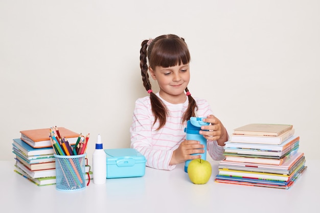 Image of smiling happy little schoolgirl with dark hair and braids sitting at table surrounded with books holding bottle with water posing during break isolated over white background