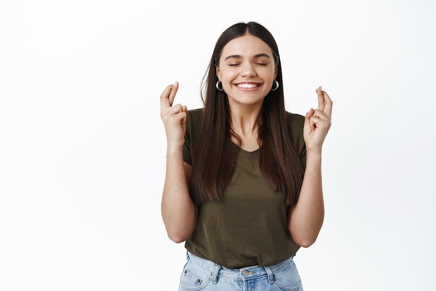 L'immagine di una ragazza sorridente e speranzosa pensa in modo positivo facendo un desiderio con gli occhi chiusi e un grande sorriso incrocia le dita per buona fortuna in piedi su sfondo bianco