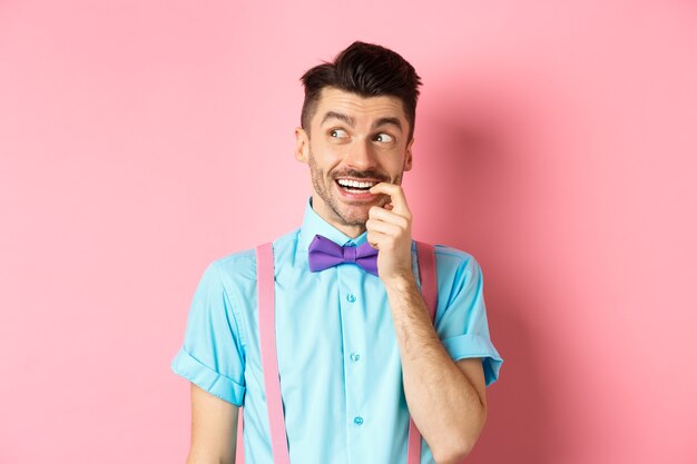 Image of smiling man making choice, looking dreamy and happy aside, biting fingernail with tempted expression, wanting something, standing over pink background.