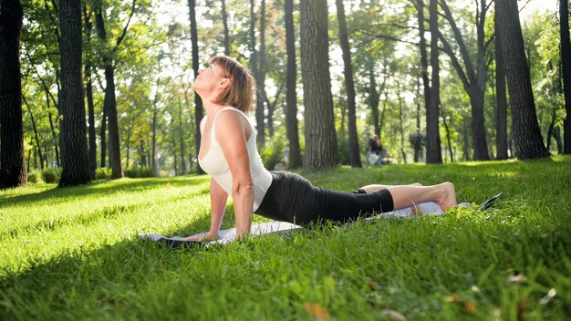 Image of smiling middle aged woman in fitness clothes doing stretching and yoga exercises. WOman meditating and doings sports on fitness mat on grass at park