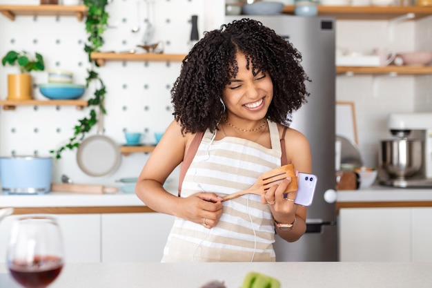 Immagine di una bella donna sorridente che indossa abiti casual usando le cuffie e ballando mentre si cucina la cena in una cucina accogliente