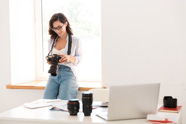 カメラとオフィスで笑顔の楽観的な若い写真家の女性の画像。