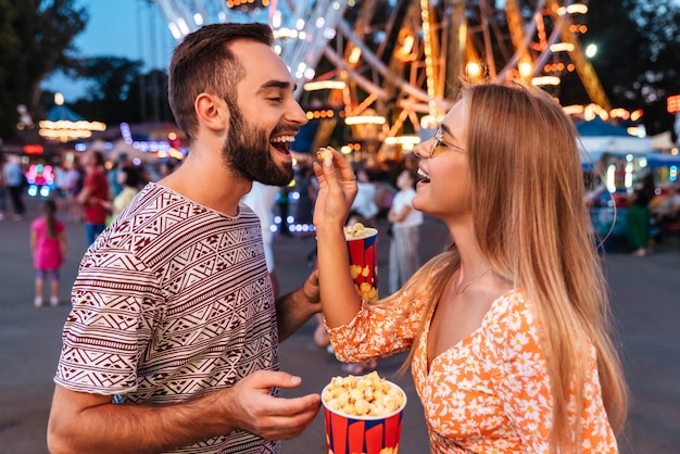 Image of a smiling positive loving couple walking outdoors in amusement park eat popcorn.