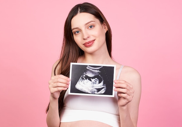 Image of a smiling pregnant cute young caucasian girl with dark hair and blue eyes, standing against a pink background. Looks straight ahead and holds an ultrasound scan.