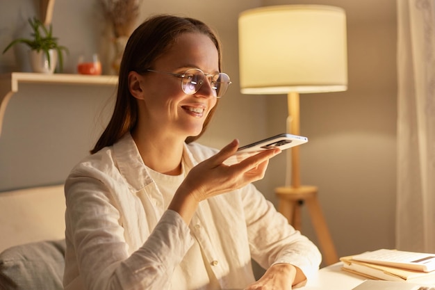 Image of smiling satisfied adorable woman with brown hair wearing white shirt sitting in evening at home holding mobile phone and recording voice message